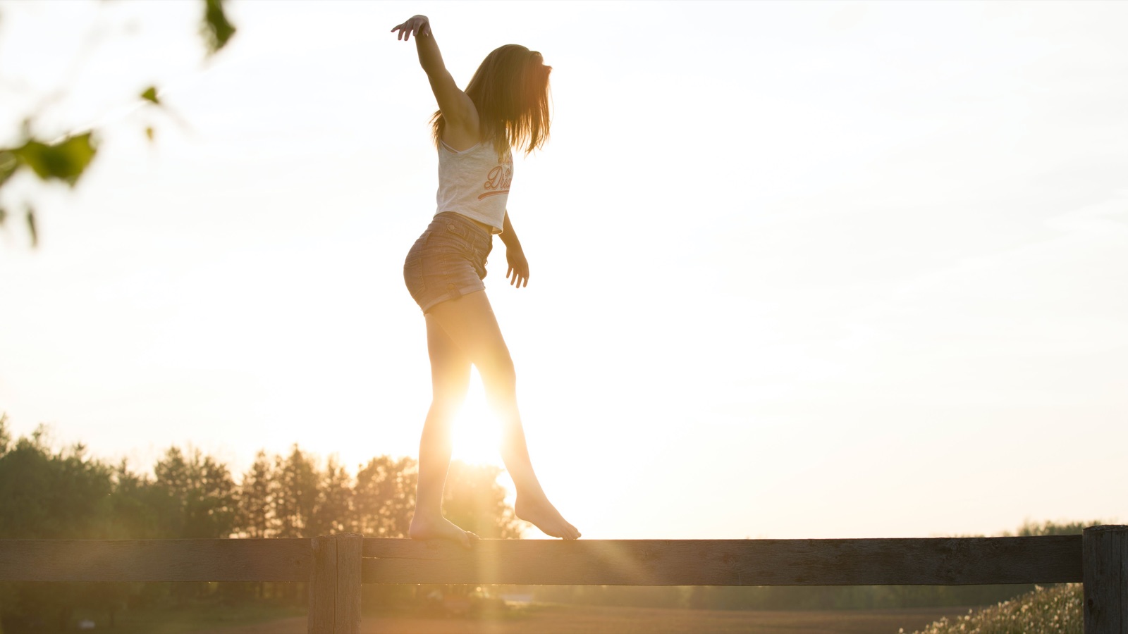Kvinde balancerer på et hegn i modlys ved solnedgang